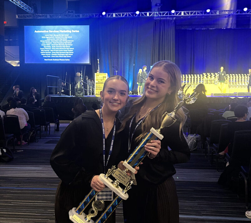Two smiling young women stand together holding a large trophy at a DECA International Competition in an auditorium with blue lighting and rows of seated people in the background.