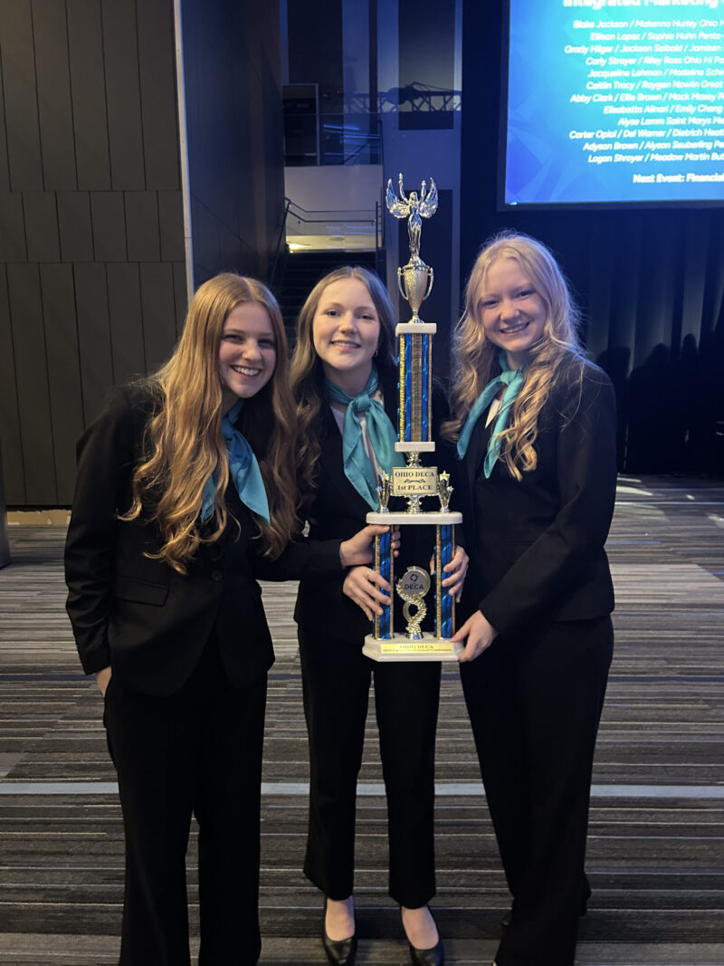 Three smiling girls in matching black suits and teal scarves stand indoors holding a large blue and gold trophy together after winning DECA State Honors. A projected blue screen with names appears in the background.