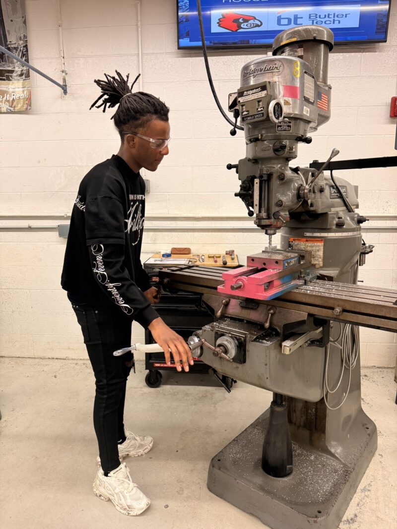 A person wearing safety glasses operates a large metal milling machine in a workshop. As part of the machining olympics, they turn a handwheel while various tools are visible on a workbench in the background.