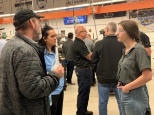 A young woman in safety glasses stands with hands in pockets, talking to a man in a cap and jacket at a busy workshop during the machining olympics. Students connect while others converse nearby; industrial machines and banners fill the background.
