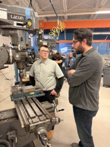 A man explains something to a student standing beside a large milling machine in a workshop during the Machining Olympics. Other people and machines are visible in the background. Both are focused on the discussion.
