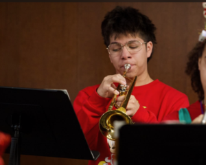 A senior wearing glasses and a red sweater plays a trumpet, standing behind a black music stand with a brown background.