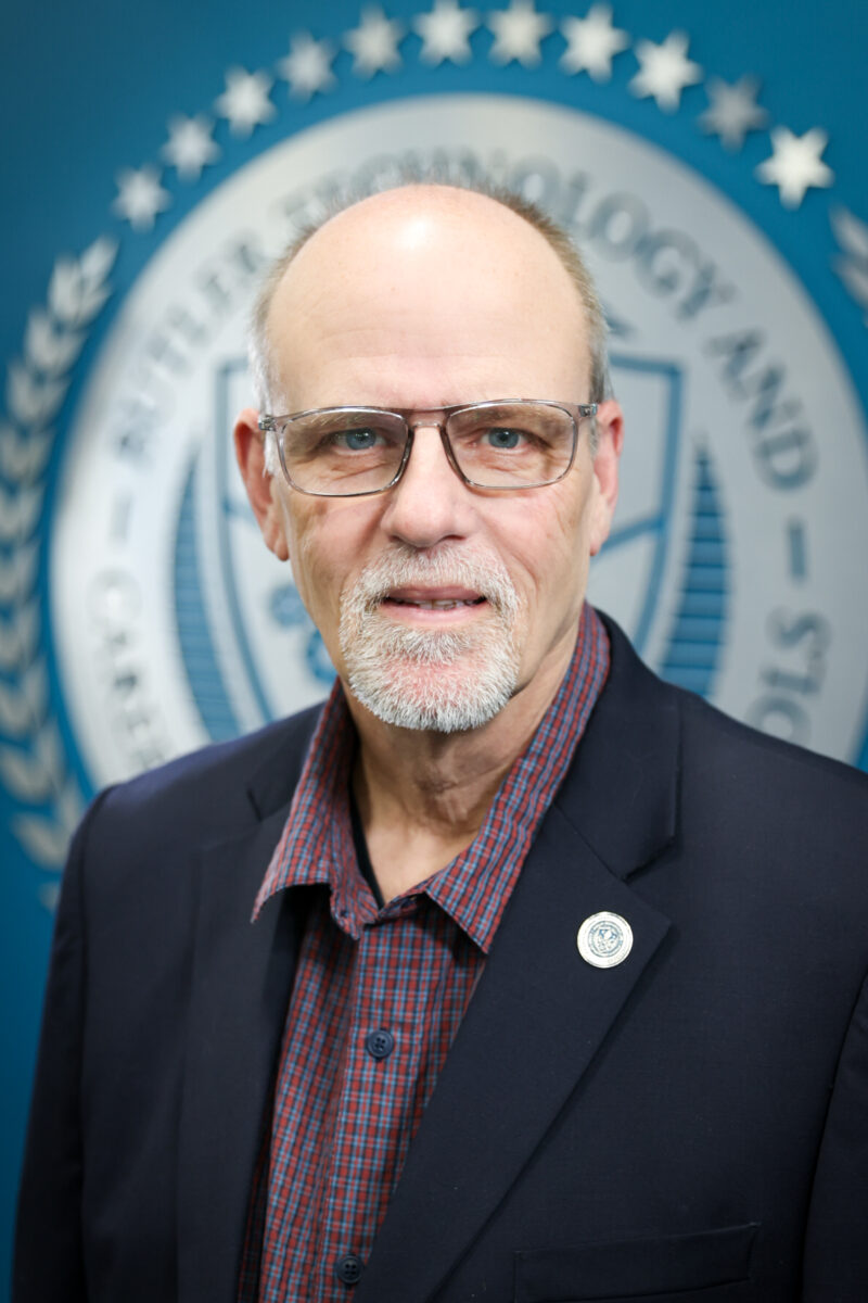 A middle-aged man with glasses, a short gray beard, and a bald head, wearing a dark blazer and plaid shirt, stands in front of a blue Board of Education backdrop featuring a circular emblem and text.