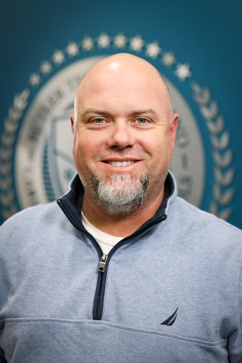 A smiling bald man with a light beard and mustache, wearing a light blue pullover, stands in front of a blurred background featuring the Board of Education's circular emblem with stars and laurel leaves.