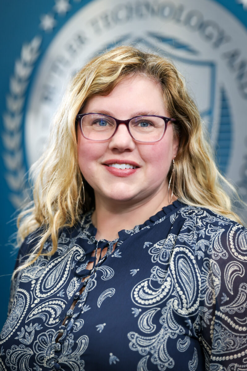 A woman with wavy blonde hair and glasses smiles at the camera, wearing a blue paisley-patterned blouse. A blurred Board of Education silver seal is visible in the background.