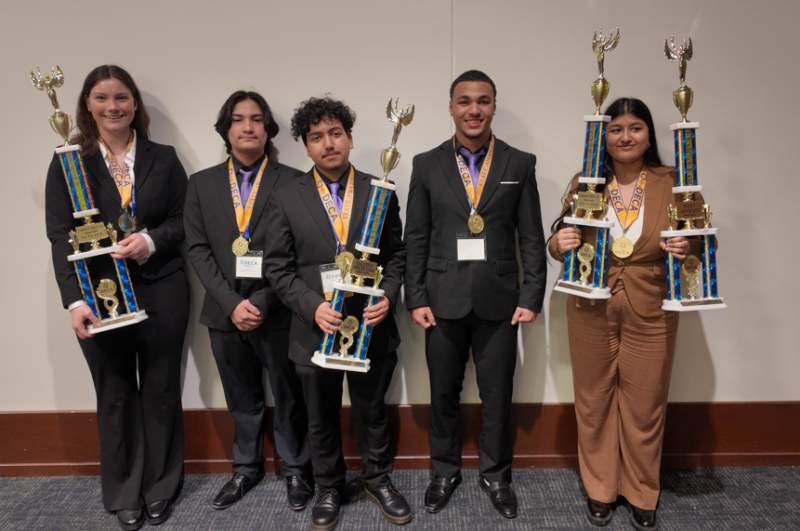 Five young adults in suits stand indoors against a wall, smiling and holding large trophies with gold medals around their necks, celebrating their DECA State Honors at an academic or competitive event.
