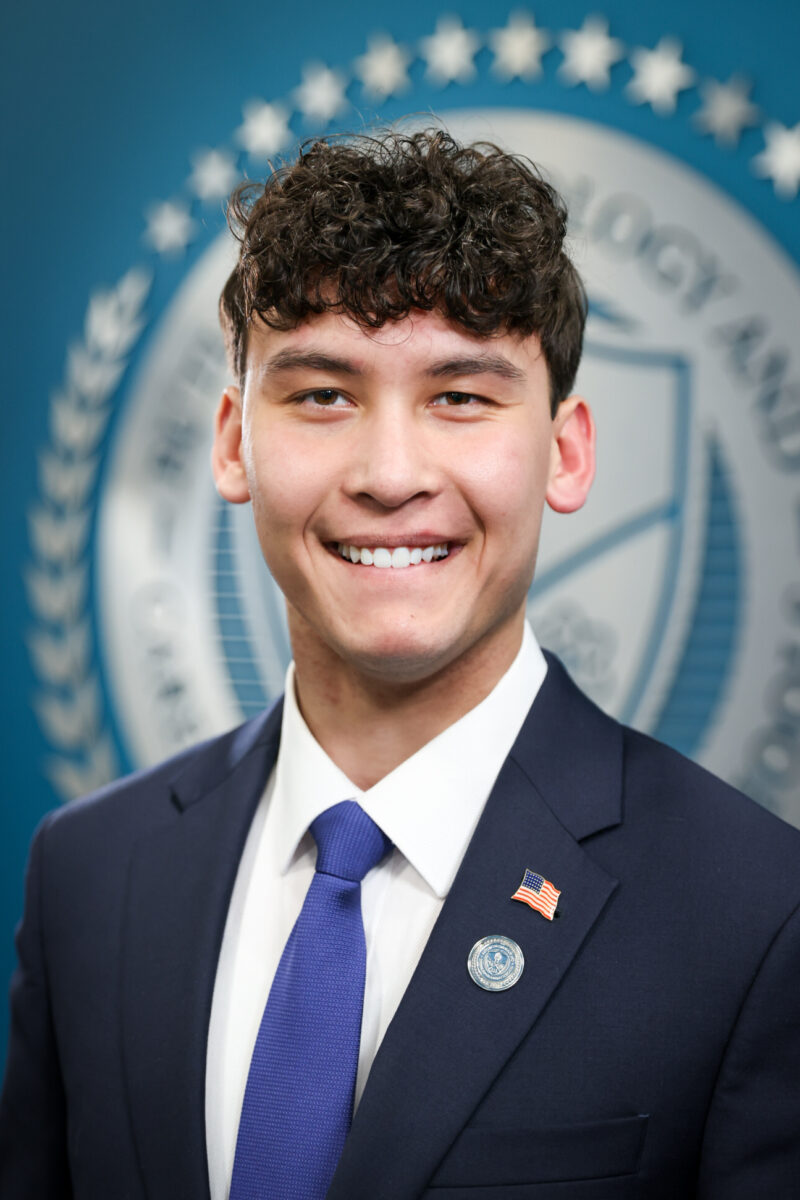 A young man with curly brown hair in a navy suit, white shirt, and blue tie smiles in front of a blurred Board of Education seal. He wears an American flag pin and a round pin on his lapel.