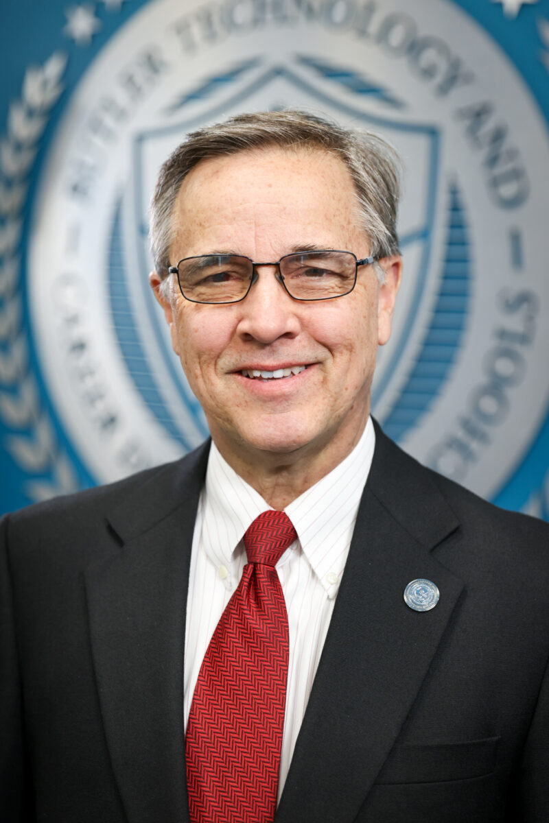 A middle-aged man in a dark suit, white shirt, and red patterned tie stands smiling in front of a blue background with a blurred Board of Education seal. He is wearing glasses and a lapel pin.