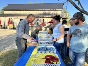 People stand at an outdoor table draped with a blue and yellow cloth, browsing items in white baskets. This Talawanda–Butler Tech FFA event highlights community culture, set against a wooden building with red umbrellas on a sunny day.