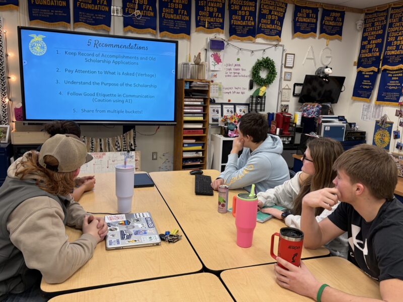 Four students sit at a classroom table, attentively watching a presentation on scholarship application tips. Talawanda–Butler Tech FFA banners hang on the wall, reflecting a culture of leadership and achievement in the room.