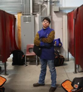 A young person wearing safety gear stands with arms crossed in an industrial workshop area, surrounded by red welding curtains and tool bags on the floor. OTC+ programs help create new pathways for both youth and seniors in skilled trades.
