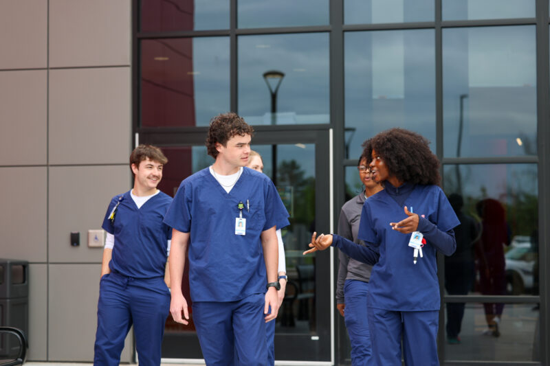 Four healthcare workers in blue scrubs walk and talk outside the modern Bioscience Center with large windows, appearing engaged and cheerful.
