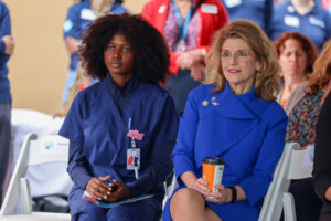 Two women sit side by side at a Ribbon Cutting event for the Bioscience Center. One wears navy blue medical scrubs and an ID badge, the other a bright blue suit and glasses, holding a coffee cup. People in matching shirts gather in the background, celebrating the future.