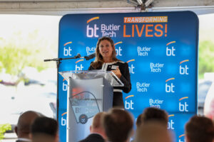 A woman speaks at a podium in front of a Butler Tech backdrop that reads "TRANSFORMING LIVES!" during an outdoor Ribbon Cutting event at the Bioscience Center, with an audience seated in the foreground and the future in focus.