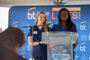 Two women in blue scrubs stand at a podium with microphones, speaking at a Ribbon Cutting event. A blue backdrop behind them displays the logos for Butler Tech and BSC Bioscience Center as they celebrate the future of bioscience.