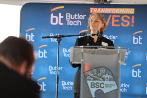 A woman speaks at a podium during a Ribbon Cutting event in front of a blue Butler Tech backdrop, which features the words "TRANSFORMING LIVES!" and "BSC Bioscience Center." A person is visible in the foreground, celebrating the future of the Bioscience Center.