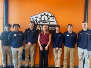 Seven people stand in front of an orange wall with a Butler Tech Aviation center logo. Six wear navy shirts and khaki pants, while one in the center, possibly a student pilot, wears a maroon blouse and black pants. They all smile at the camera.