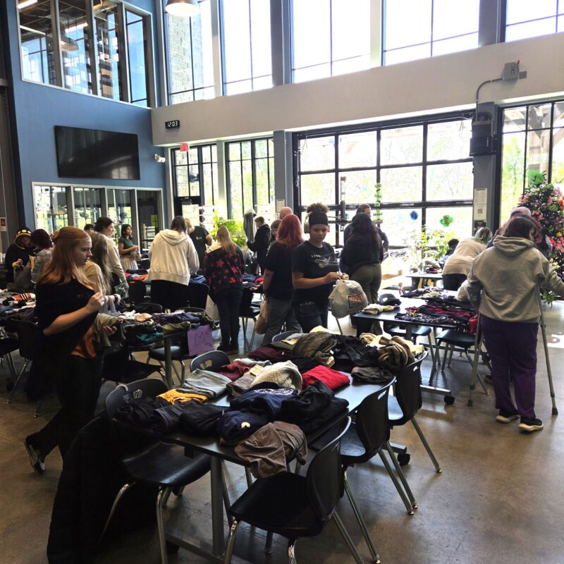 People browse and sort through tables filled with clothes at an indoor clothing swap event in a bright, spacious room with large windows, promoting sustainability as they shop, chat, and organize items.