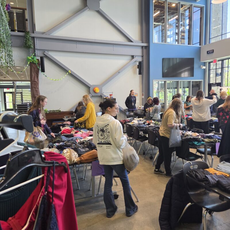 People browse tables filled with clothes and accessories in a spacious, modern indoor area with large windows. Some are examining items, while others carry bags, suggesting a sustainable clothing swap inspired by Earth Day.