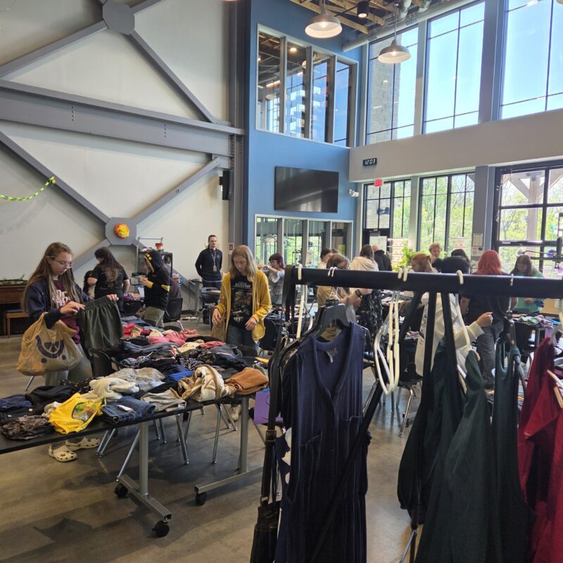 People browse clothing racks and tables filled with clothes at an indoor clothing swap event. Large windows let in natural light, while several people examine garments in support of sustainability and Earth Day in a spacious, modern room.
