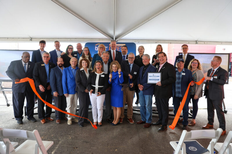 A group of around 25 people pose under a tent at the Bioscience Center ribbon cutting ceremony, smiling and holding a large orange ribbon and oversized scissors. Some hold a certificate; chairs are visible in the foreground, marking an exciting future.