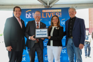 Four adults in business attire smile and pose under a tent at the Bioscience Center ribbon cutting. Two hold a framed certificate. Behind them, a blue backdrop reads "bt Butler Tech" and "Transforming Lives!" in orange and white, celebrating the future.
