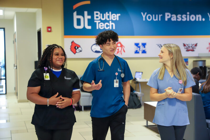 Three students in medical scrubs walk and chat inside a bright hallway at the Butler Tech Bioscience Center. Two females and one male wear badges and stethoscopes, smiling in front of a blue Butler Tech sign on the wall.