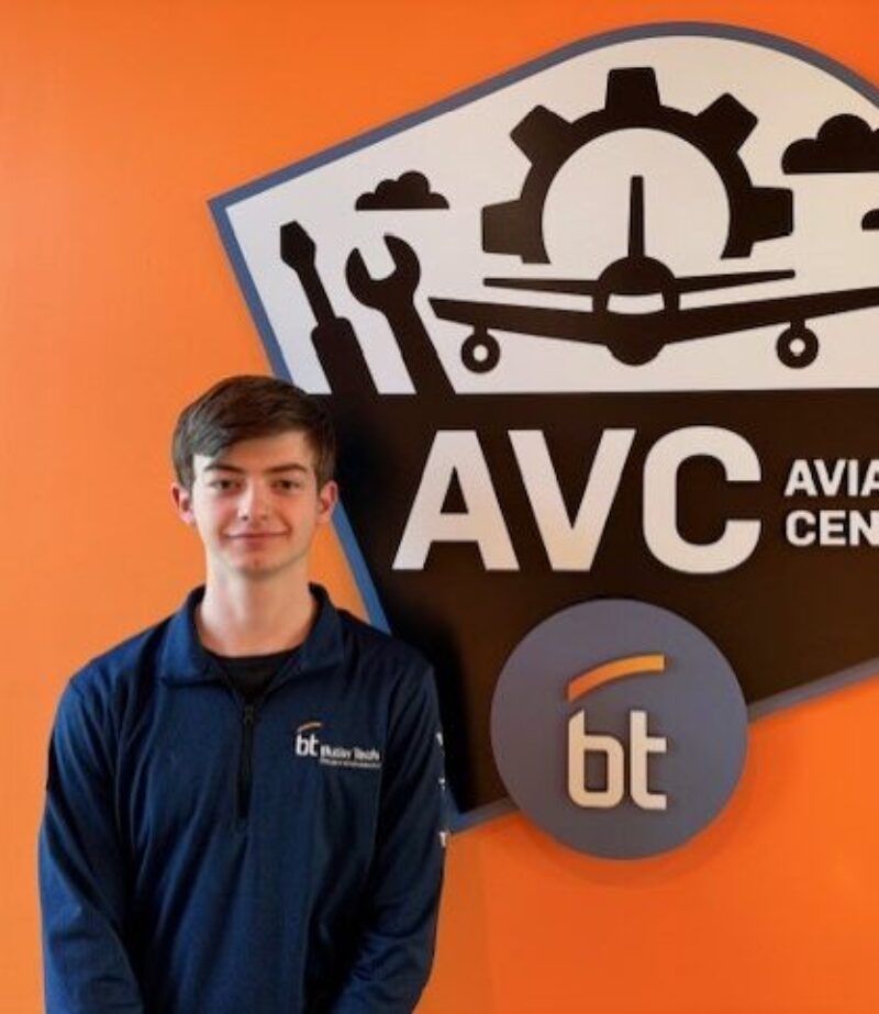 A student pilot stands in front of an orange wall with a sign that reads 
