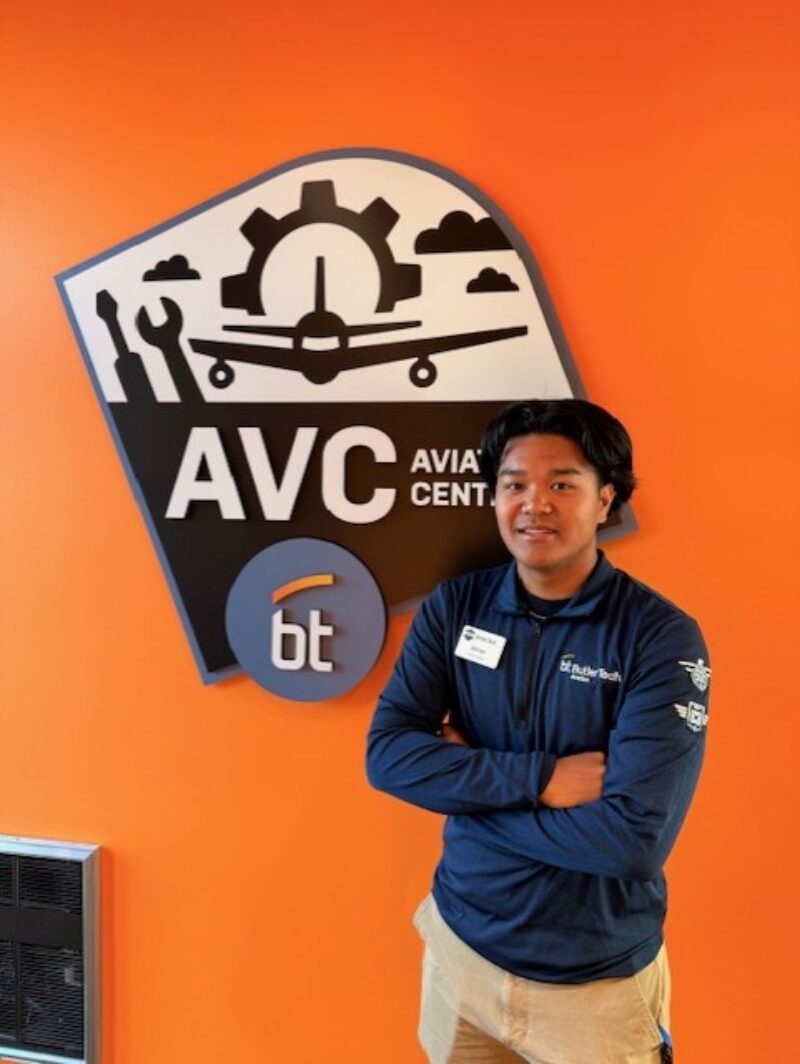 A man with dark hair stands with arms crossed, smiling in front of an orange wall featuring a large 