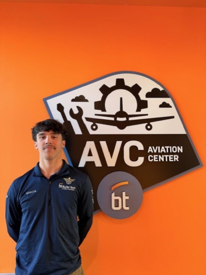 A young student pilot in a dark blue shirt stands in front of an orange wall with a sign that reads 