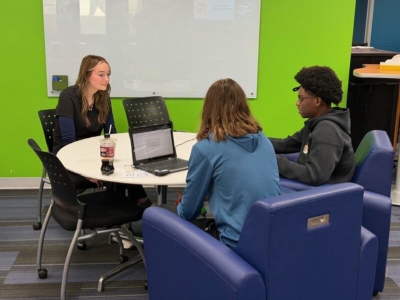 Three Cosmetology Students from Butler Tech sit around a small table with a laptop and drinks in a modern classroom, engaged in discussion. A whiteboard and bright green wall are visible in the background.