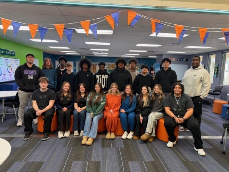 A group of 19 Butler Tech cosmetology students pose together in a brightly lit classroom, with orange and blue flags hanging above. Seven sit in the front row, and twelve stand behind them, all smiling at the camera.