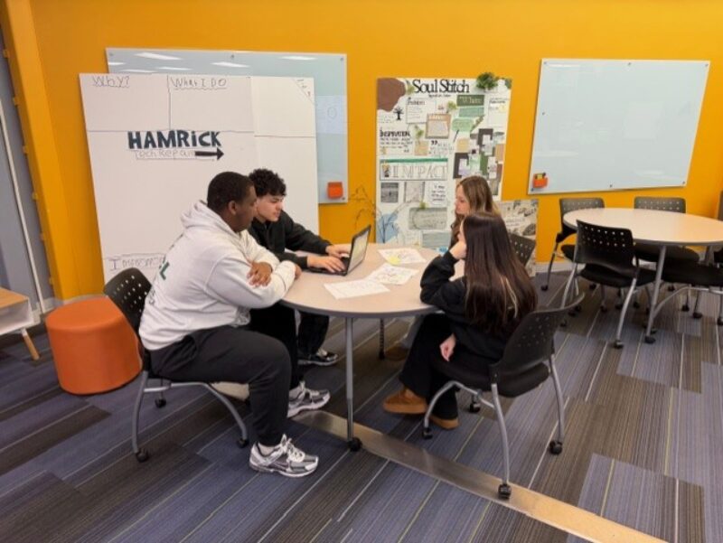 Four Butler Tech cosmetology students sit around a circular table in a classroom with yellow walls, collaborating and looking at papers and a laptop. Whiteboards, posters, and Middletown Marketing materials are visible in the background.