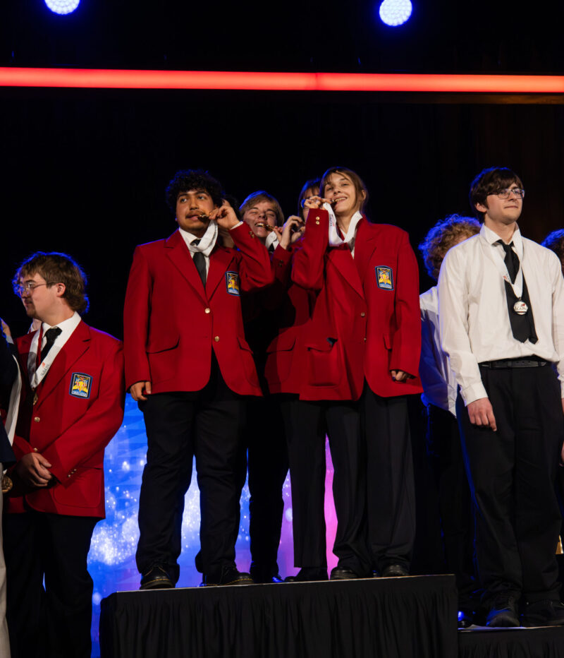 A group of Butler Tech students in red blazers and medals stand on a stage platform, celebrating and posing proudly at the State Conference, while others stand and smile beside them under bright stage lights.