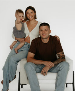 A smiling woman holds a baby on her hip while standing next to Ricky Davis, featured in our Alumni Spotlight for Industrial Maintenance Technology. All three pose together in casual outfits against a plain white background.