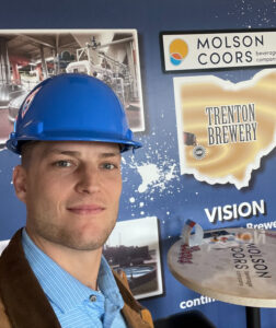Alumni Spotlight: Ricky Davis, a graduate of Industrial Maintenance Technology, smiles for a selfie wearing a blue hard hat and brown jacket in front of the Molson Coors Trenton Brewery sign, with company materials and industrial equipment in the background.