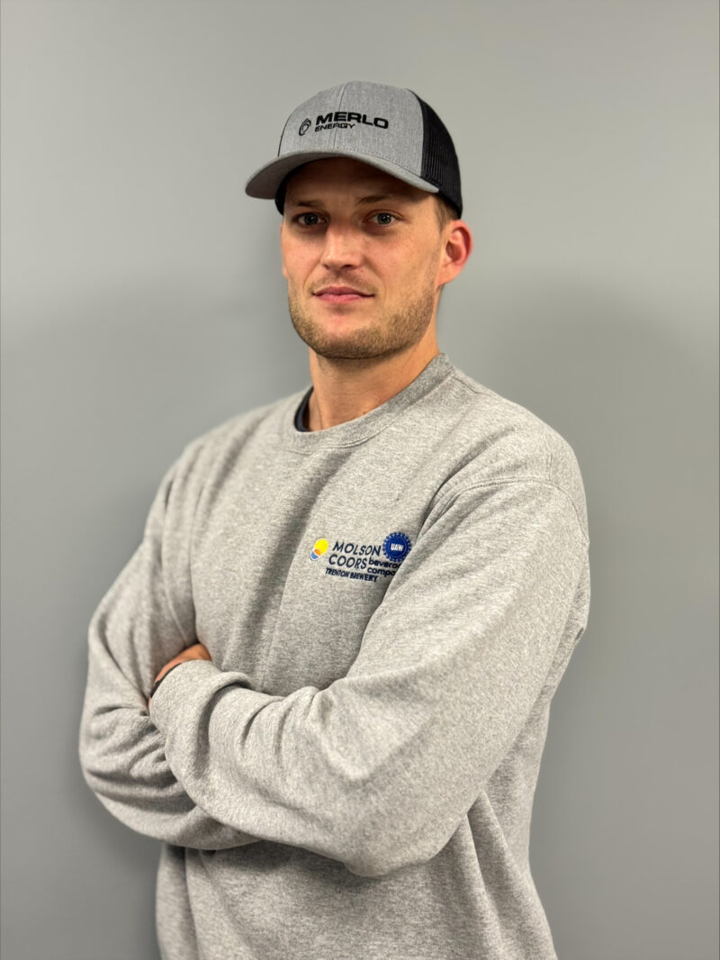 A man wearing a gray Merlo cap and a gray Molson Coors sweatshirt stands with his arms crossed in front of a plain gray wall for an Alumni Spotlight in Industrial Maintenance Technology.