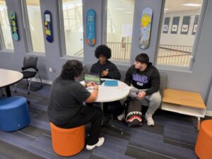 Three students, including Cosmetology Students from Butler Tech, sit around a round table in a modern school lounge, chatting and working on a laptop. Skateboard art decorates the walls and large windows look into a busy hallway.
