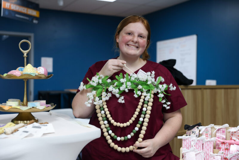 A smiling person with red hair in maroon scrubs holds up a large, decorative garland of wooden beads and white flowers at a table of crafts and pastries displayed for the Entrepreneurship Fair.