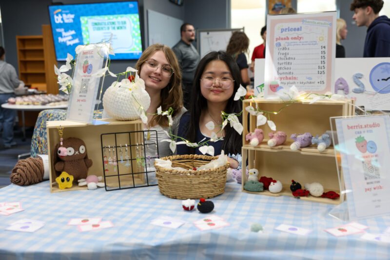 Two young women sit behind a booth at the Entrepreneurship Fair, displaying handmade crochet plushies—cows, frogs, and chicks. The table features a blue checkered cloth, signs, a price list, and small string lights showcasing student businesses.