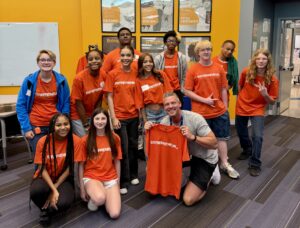 A group of teens and two adults pose indoors, all wearing bright orange "ENTREPRENEUR" shirts except one adult holding up a shirt. Smiling, they stand and kneel in front of yellow and gray walls—Future Innovators at a Summer Camp for entrepreneurship.
