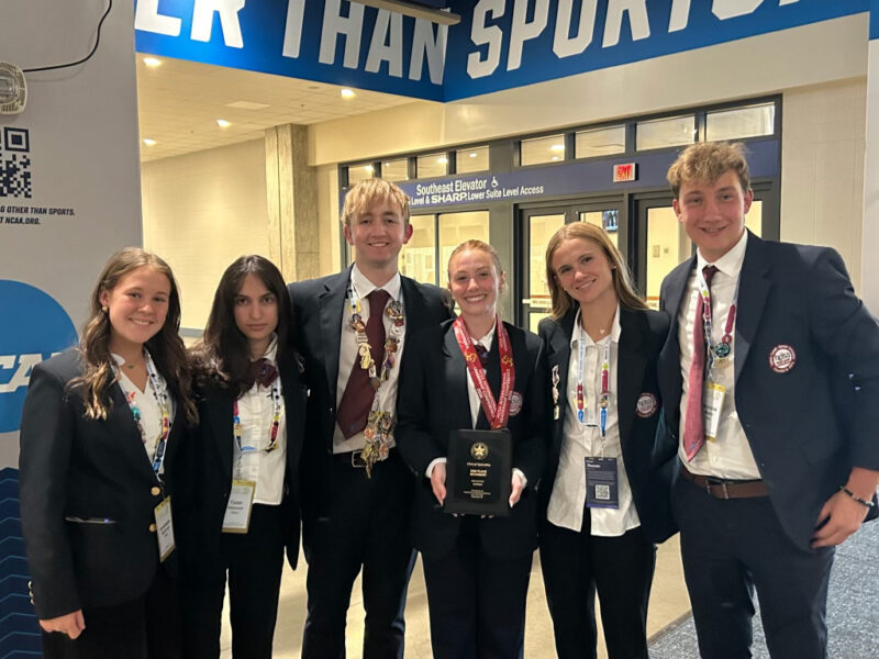 Six smiling students in formal attire stand together indoors, some wearing medals and badges at the HOSA Leadership Conference. The center student holds a black award plaque beneath a large blue sign. The Butler Tech group appears happy and proud.
