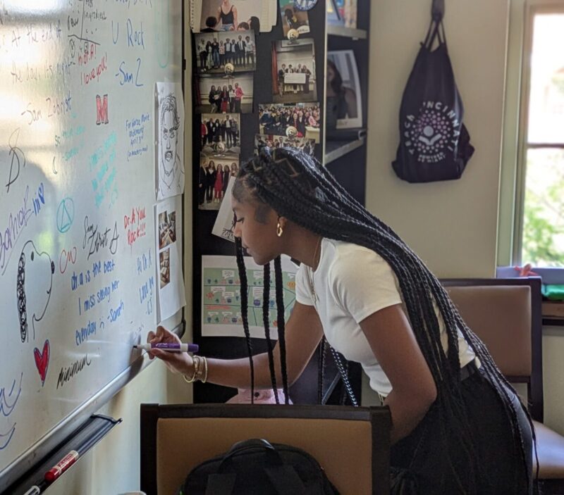 A young woman with long braids writes on a whiteboard filled with colorful messages, drawings, and notes in a bright classroom or office setting. Photos and bags are visible on the wall nearby.