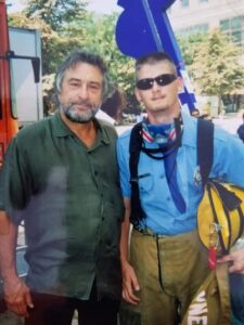 Two men pose outdoors: Jim Miller in a green shirt with gray hair and beard, and another in firefighter gear with sunglasses, holding a yellow helmet. Trees and buildings frame their Butler Tech journey in the background.