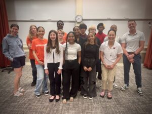 A group of thirteen teens and three adults stand together, smiling in a classroom. Some wear matching orange “Entrepreneur” shirts, while others wear casual or business casual outfits. A clock and whiteboard are visible in the background.
