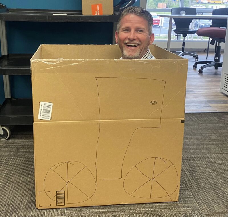 A smiling man, possibly Jim Miller from Butler Tech, sits inside a large cardboard box with a bicycle drawn on the side, making it look like he is riding it—creating a playful 