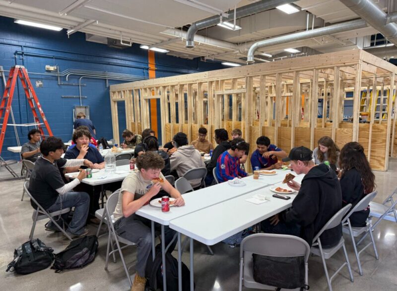 A group of construction students sit at folding tables eating pizza and drinking soda in a workshop with an unfinished wooden structure in the background. Ladders and backpacks are visible, reflecting a hands-on construction education environment.