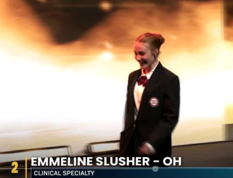 A young woman in a formal dark blazer walks onstage, smiling. The text reads: 
