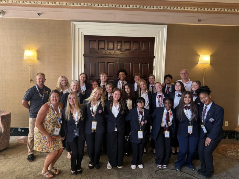 A group of young adults, most wearing dark blazers and medals, pose together indoors at the International HOSA Leadership Conference in front of large wooden doors. Two adults, representing Butler Tech, stand at the sides, smiling for the camera.
