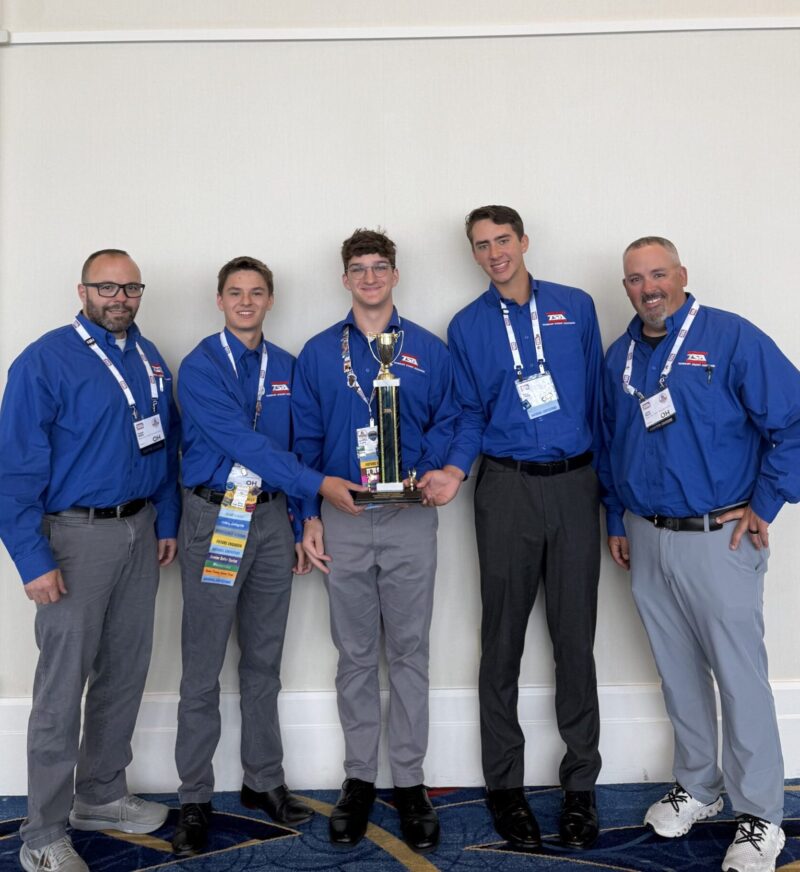 Five men in matching blue shirts and gray pants stand indoors, smiling and posing with a trophy from the TSA Championships. Four of the BT Students wear name badges on lanyards, standing proudly against a plain white wall.